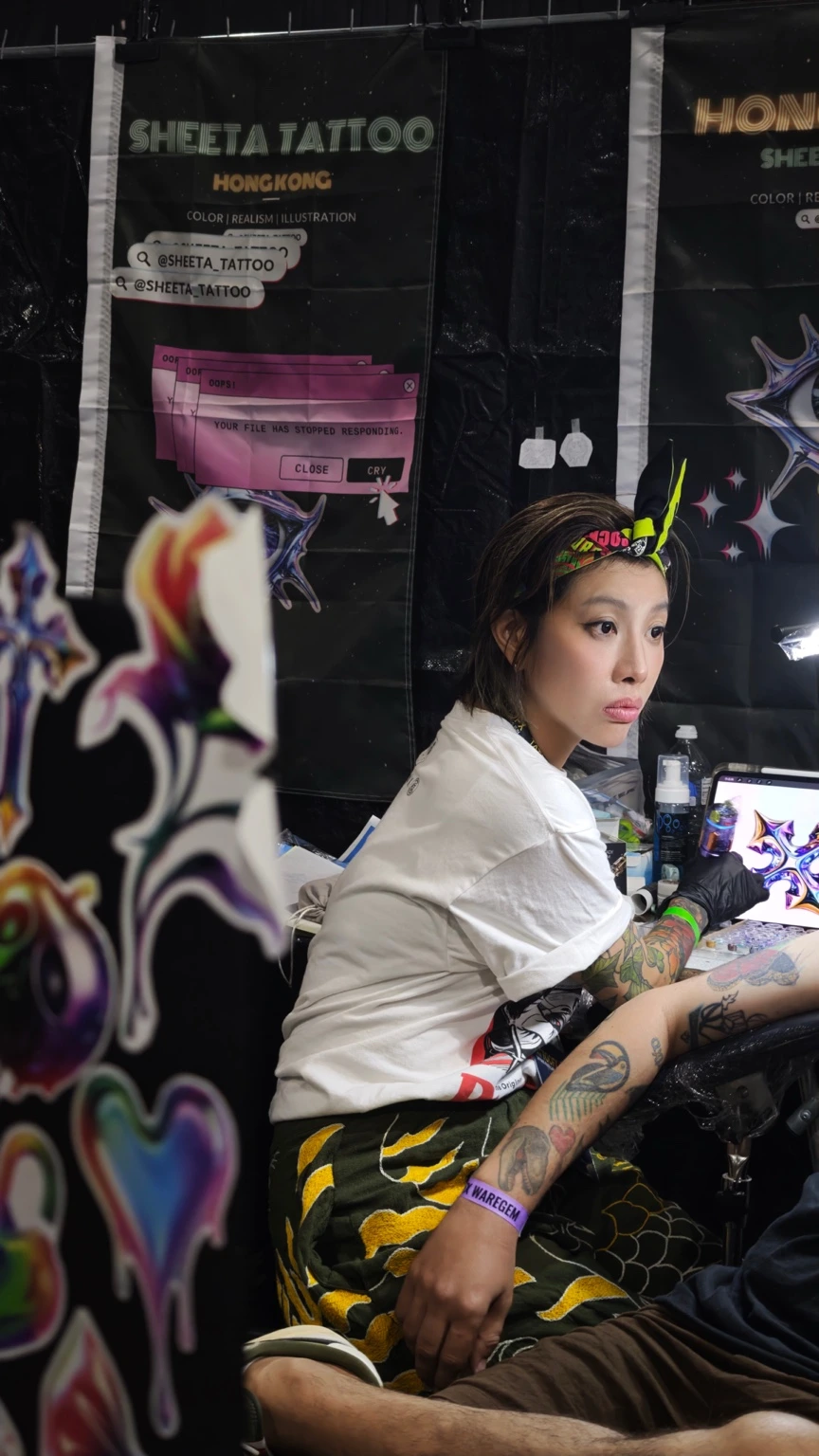 香港紋身師 Sheeta 在紋身展攤位工作，戴著手套，身旁展示幻彩金屬紋身設計。